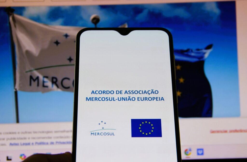 Tela de celular exibindo o acordo Mercosul–União Europeia com bandeiras do Mercosul e da União Europeia ao fundo.